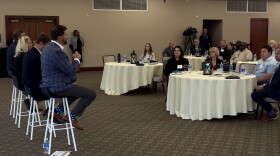 Four individuals sit on stools in front of people sitting at several round tables with white tablecloths. The person closest to the camera, on a stool,  is holding a microphone. 