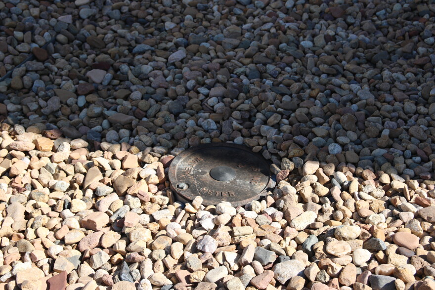 A metal water meter sits in the ground surrounded by tan landscaping rocks.