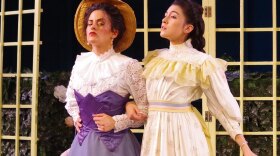 Two women in Victorian dresses stand arm in arm, scoffing at one another