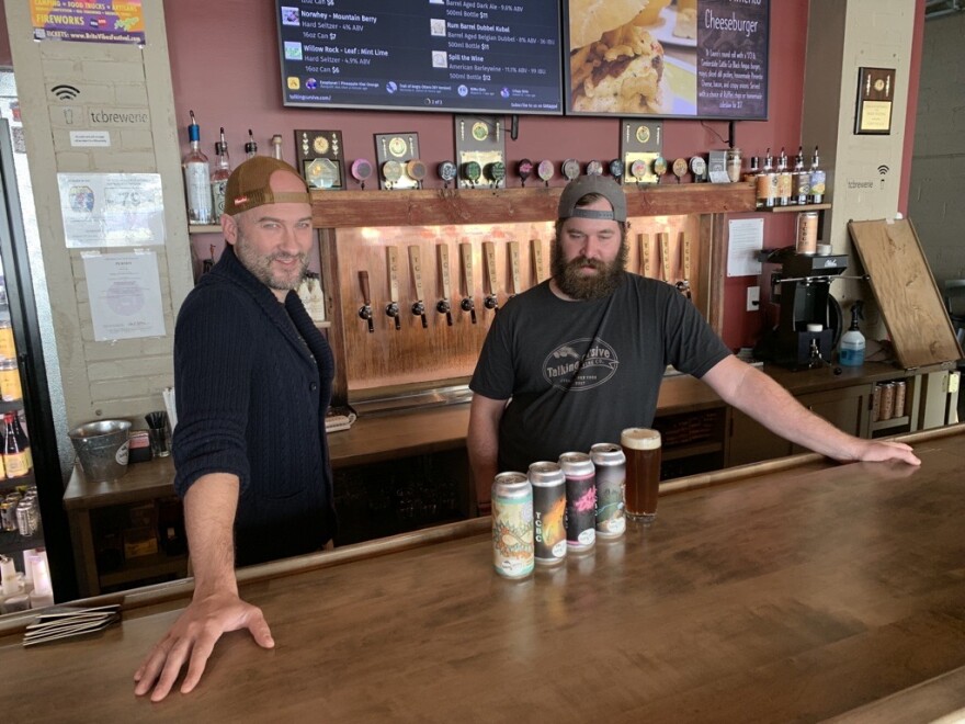 Two men stand behind a bar counter with four beer cans placed in front of them.