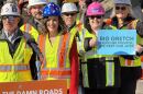Governor Gretchen Whitmer stands among a group of construction workers in a customized blue hard hat with stars.  Whitmer and the construction workers are all wearing neon yellow safety vests. A female construction worker is holding a sign that says, "Big Gretch keeps her promise, we keep our jobs."
