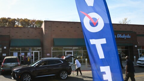 Outdoor photo showing a strip center in background. In foreground is a blue vertical flag waving in the wind. It reads "VOTE." A person at right can be seen walking toward the strip center. A car parked in front has it's lift gate open and a woman is about to place a walker into the back of it.
