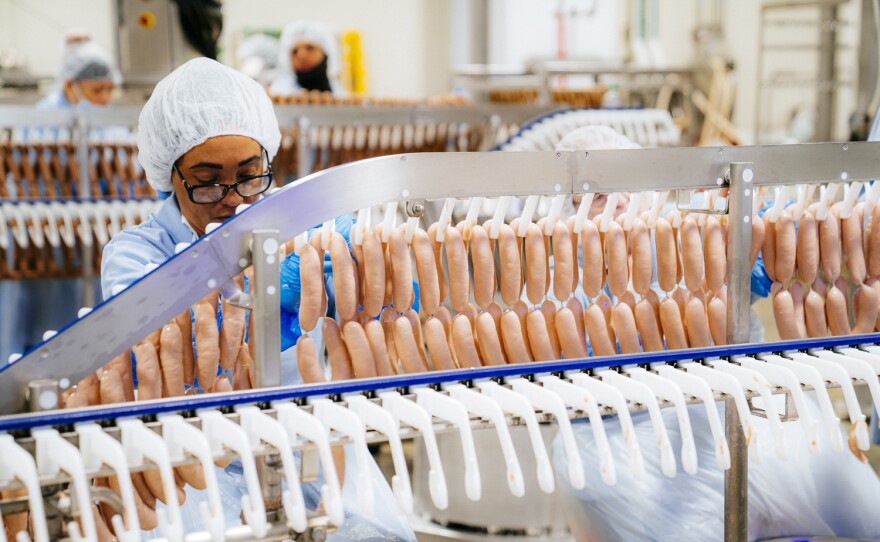 A worker wearing a hair net, glasses, white coat, and blue rubber gloves, working on a hot dog line