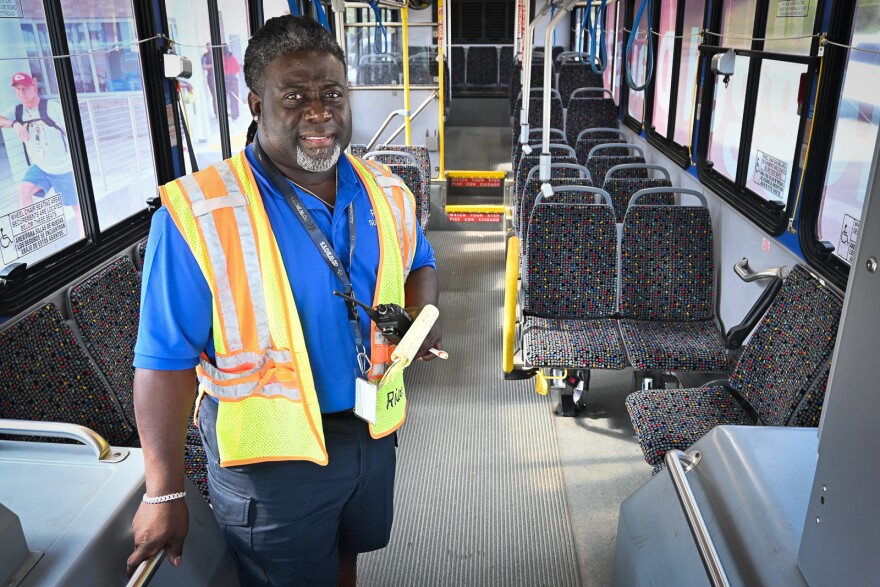 A man wearing a bright yellow safety vest holds a walkie-talkie and yellow writing pad while standing in the aisle of a city passenger bus.