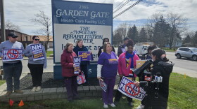 A group of 10 people stands in front of a large nursing home sign, holding up personal signs of their own calling for improved labor practices. Several vehicles drive by on an adjacent street.