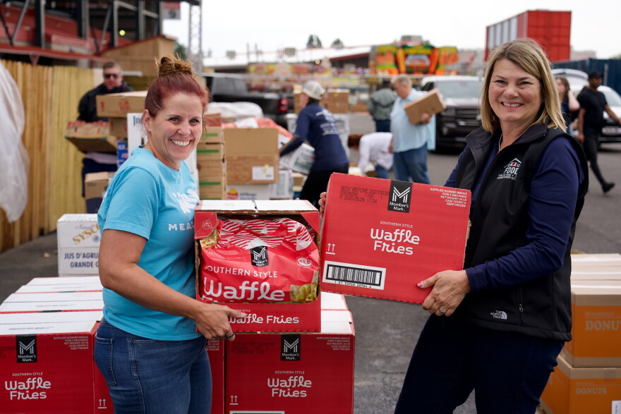 Two women, one with red hair in a bun on the left and First Lady Jennie Gordon on the right, smile while holding boxes of waffle fries. In the background, other people move boxes of food at a mobile food pantry.