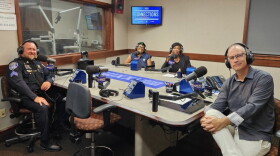 Four smiling people sit around a table in a radio talk studio: a man front left has short light brown hair and a light brown mustache and beard and is wearing a police uniform; a woman back left has short blue hair and is wearing glasses and a blue top; a woman back center has long black hair and is wearing a black top and blazer; a man at right has very short dark hair and is wearing glasses, a grey button-down shirt, and white linen pants