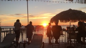 People under a tiki umbrella and leaning against a railing to watch a sunset