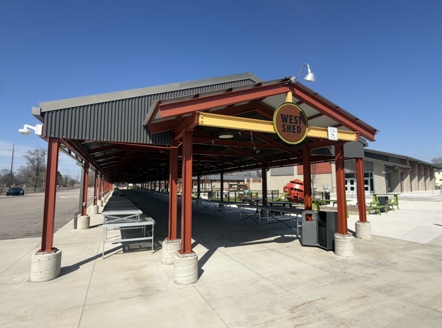 A large metal structure can be seen that looks like a long roof covering a sidewalk. It is painted red and says "west shed". Underneath it, folding tables are lined up.