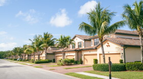 A row of side by side, tan two story houses with big garage doors in a Florida neighborhood with palm trees and bushes in front of the houses.  