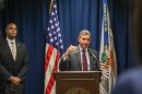 A man in a suit and tie speaks at a podium, with a U.S. flag and another flag behind him. The second flag has a banner that reads "Southern District of Ohio." A second man in a suit and tie stands to the side, hands folded. 