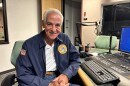Older man with jacket and jeans smiles to camera. Sits next to microphone in a studio. 