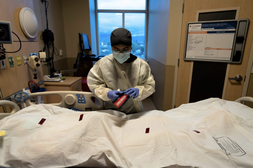  A nurse hovers over a body bag and holds a "Covid Patient" label in their hands, in a hospital room.