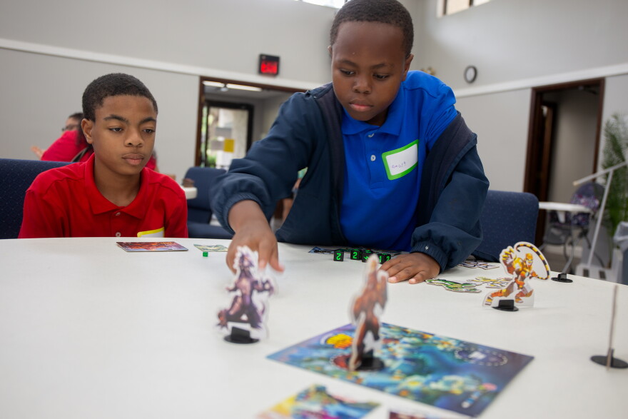 Two young boys, one in a blue shirt and one in a red, move monster tokens around on a game board.