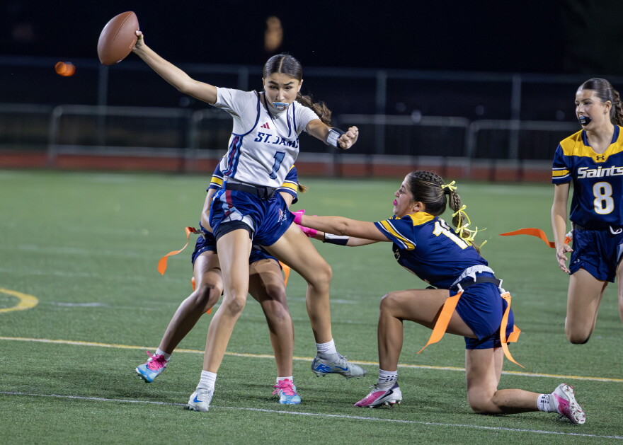 Quarterback Emme Albright is rushing and extending the ball beyond the end line. Two St. Thomas players are attempting to grab her flag. One is kneeling on the ground and reaching for Albright's flag belt.