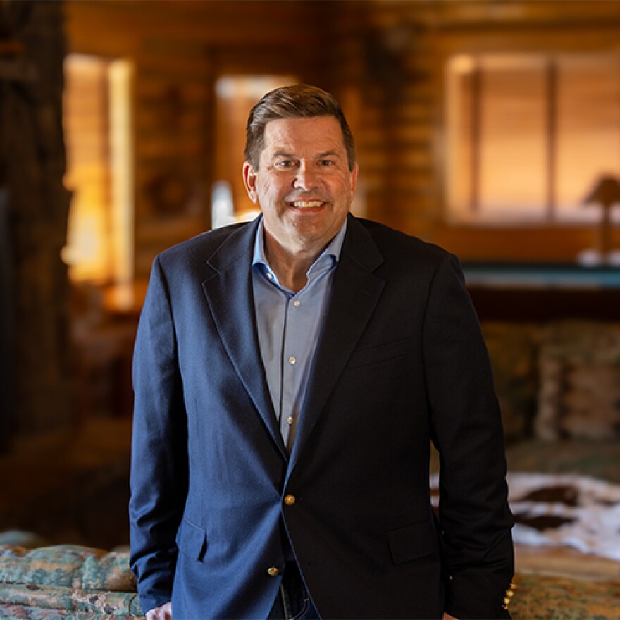 A man wearing a dark blazer smiles for the camera. He’s standing inside a log cabin room.