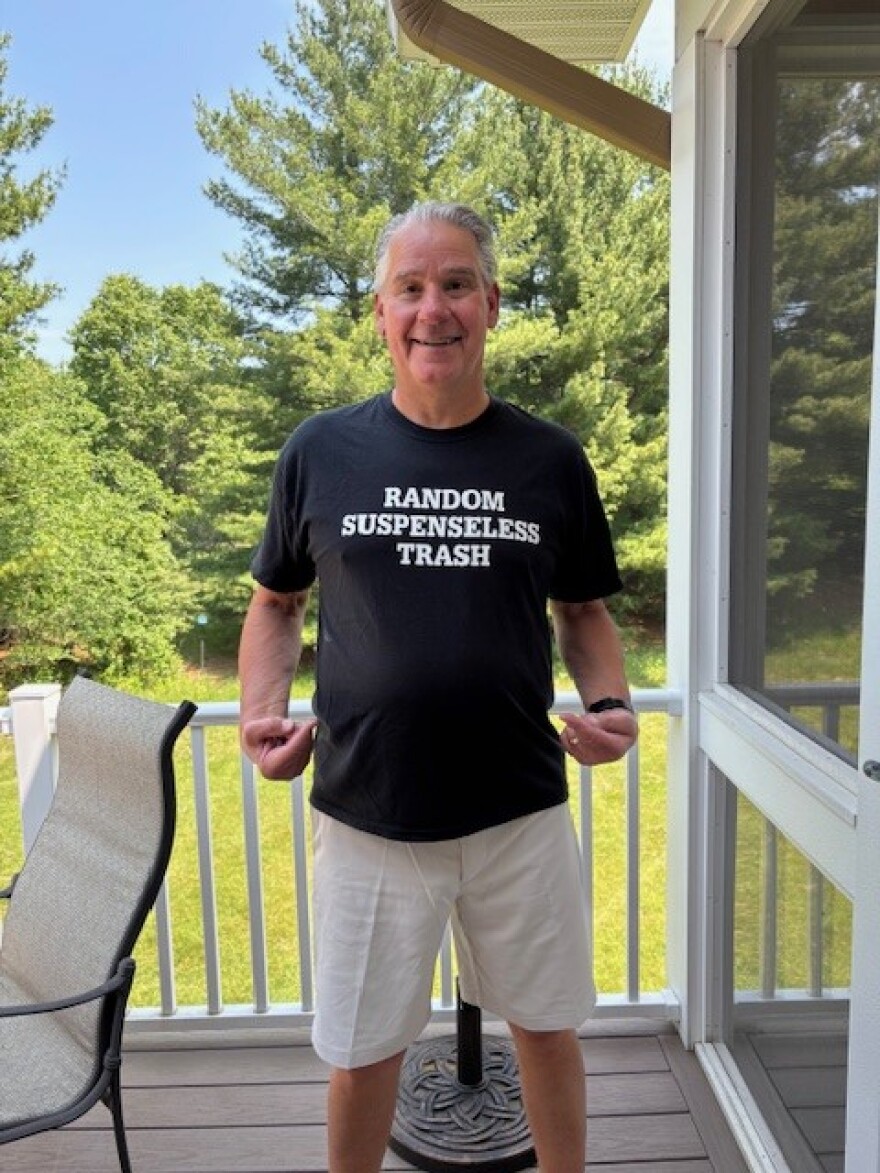An author stands on a porch with a tee shirt that shows the wording of a critique of his latest book, random suspenseless trash.