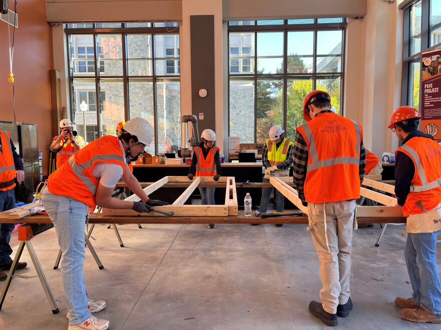 Inside a room with large windows, sunlight shines through as students wearing bright orange safety helmets and vests hammer wooden frames, which are resting on carpentry work tables.