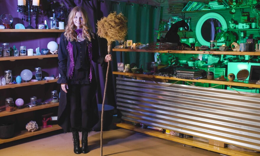 Woman dressed as witch stands with broom in front of witchy background of "magic" items.