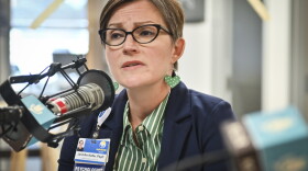 A woman wearing a blue jacket sits inside a radio studio at a microphone. She appears to be talking.