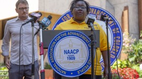 A woman in a yellow shirt stands in front of a NAACP podium with press mics positioned in front of her. 