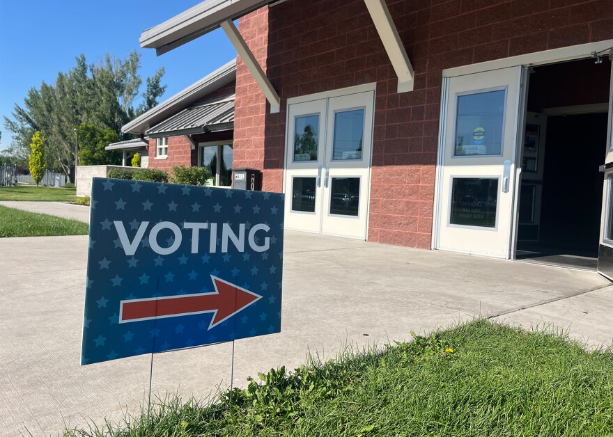 A blue voting yard sign with a red arrow pointing to a brick building stands on green grass. Green deciduous trees stand tall in the background.