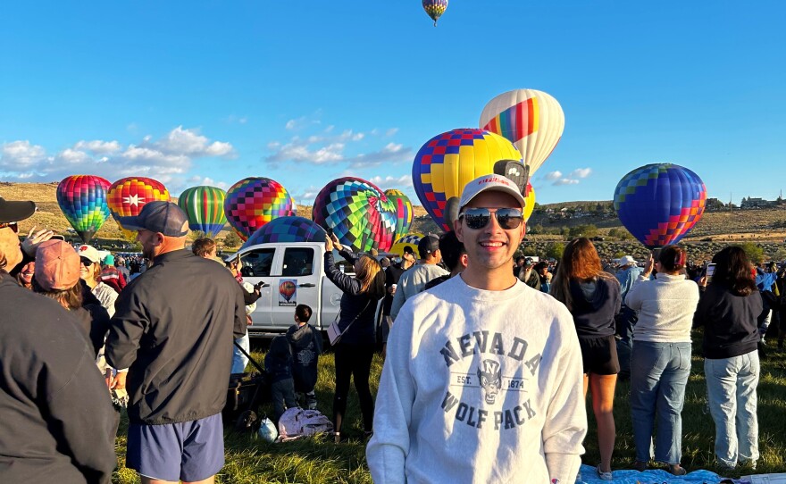 A man standing outside while looking toward the camera and smiling. In the distance are hot air balloons on the ground and in the sky.