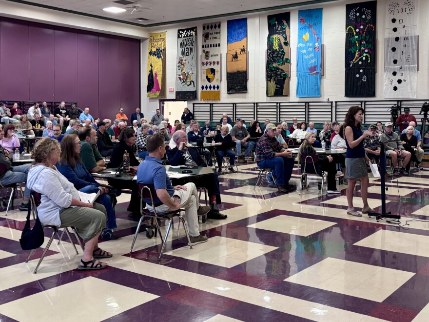 About 150 people in chairs listen to a woman standing at a podium inside an elementary school gymnasium, with musical posters on the wall.