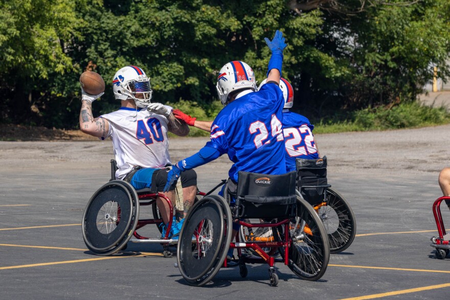 Three people in sports wheelchairs face each other. Two are in blue Buffalo Bills jerseys facing Matt Daniels in a white jersey, who is throwing a football. They are outside in a parking lot with trees behind them.