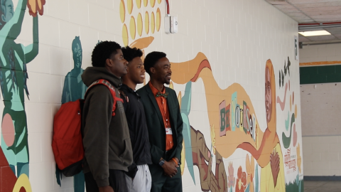Two student artists pose on Tuesday, March 31, 2026, with Eastside Principal Leroy Williams in front of the section of the mural they completed. They painted themselves playing football. (Zoe Kahn/WUFT News) 