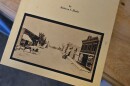 A small, tan-colored book sits on a wooden table with a medium finish. The front cover of the book bears the words "A Tribute to Johnstown by Rebecca S. Healy" in black text. A black-and-white photo features Johnstown's early downtown area. Horse-drawn carriages travel on a dirt road lined with shops.