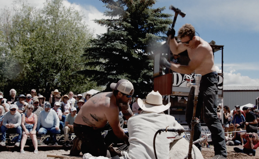 three men participate in a mining competition in Creede, Colorado. One shirtless man wields a large pickaxe