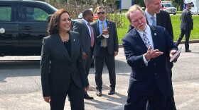 A University at Buffalo administrator and Vice President Kamala Harris are standing on a paved road. The administrator is gesturing toward something out of frame, and Harris is smiling. A Black SUV and several people in suits are behind them. The sky is partly cloudy and bright blue. Several trees and plants are in the background. 