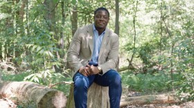 A Black man in jeans and a blazer sits on a tree stump in a wooded area.