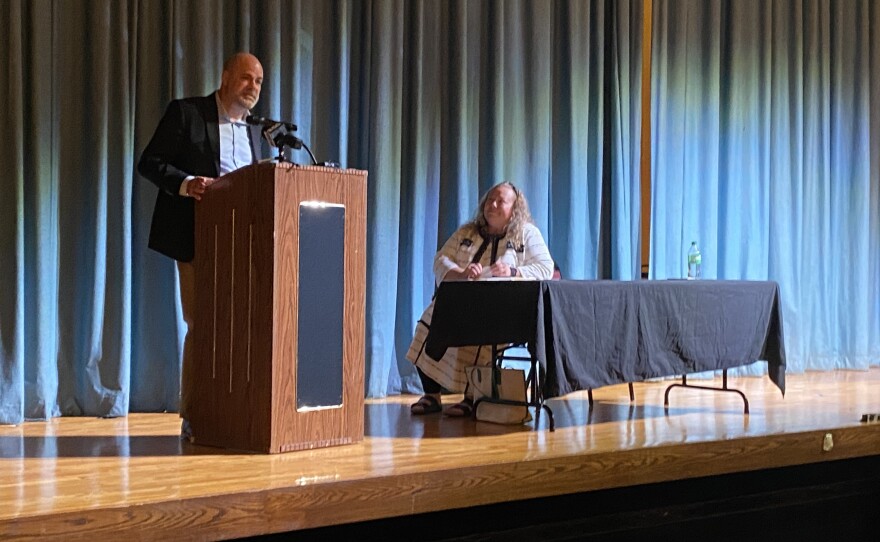 Paul Callanan stands behind a podium on stage as he gives his presentation at South Division High School. The moderator is sitting behind a table next to him.