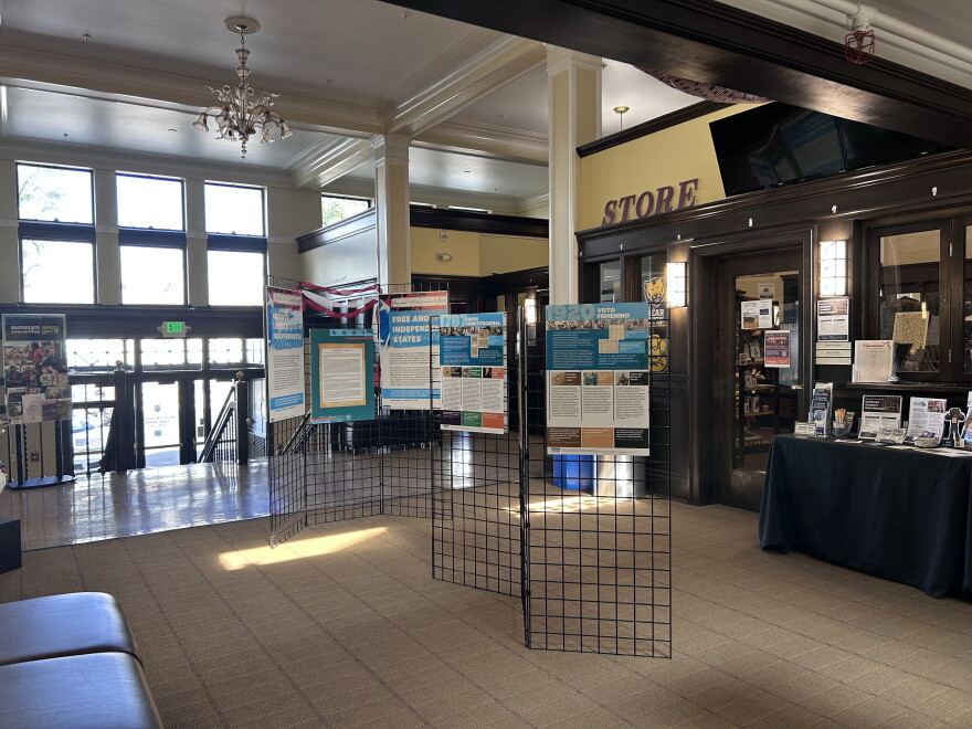 A lobby area has black metal trifold stands that have posters on them with historical information. Behind them is a wooden door with the word "STORE" above it. There's stairs leading down to the front doors of the museum. 
