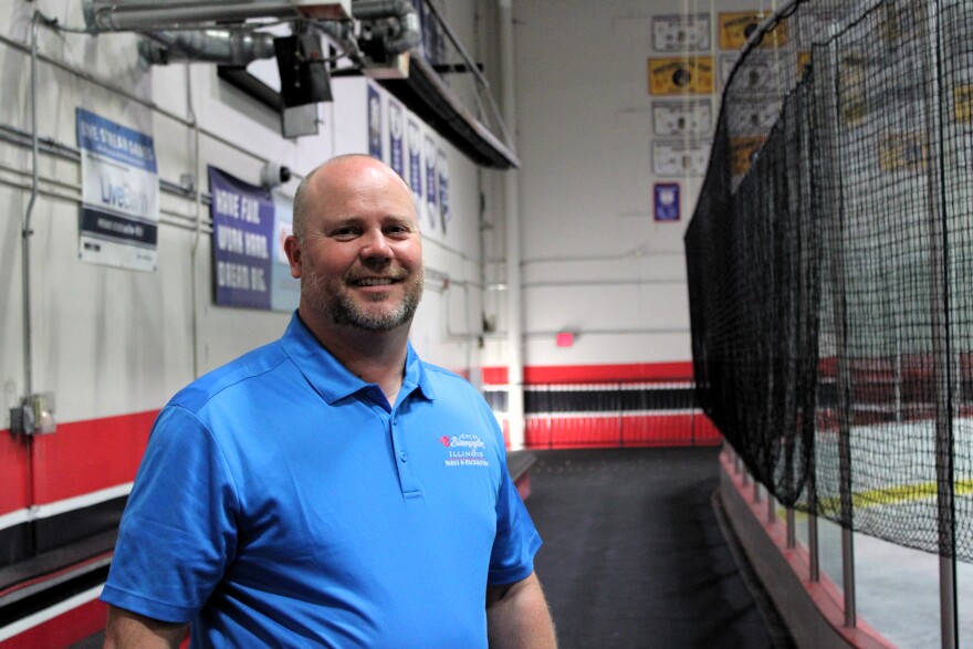A man in a blue polo shirt stands smiling inside an indoor sports facility next to a rink with protective netting. Signs and banners hang on the walls in the background.