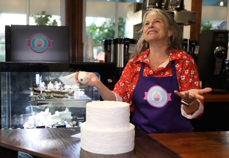 A grey haired white woman wearing an apron gestures as she decorates a two tiered cake.