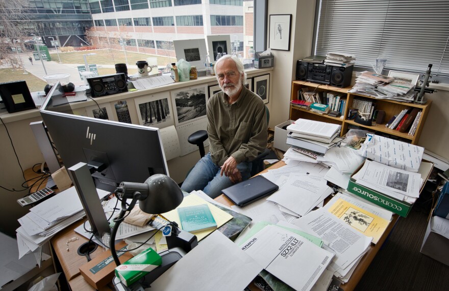 A man sits at a desk covered in stacks of papers.