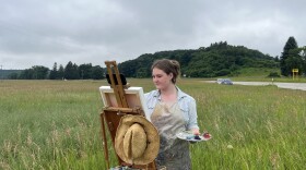 Elsa Carter painting in a field: easel, hat, and pallette in the foreground
