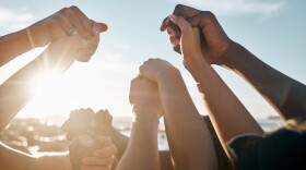 A diverse group of people holding hands in the sunshine. Only their forearms and hands are visible.