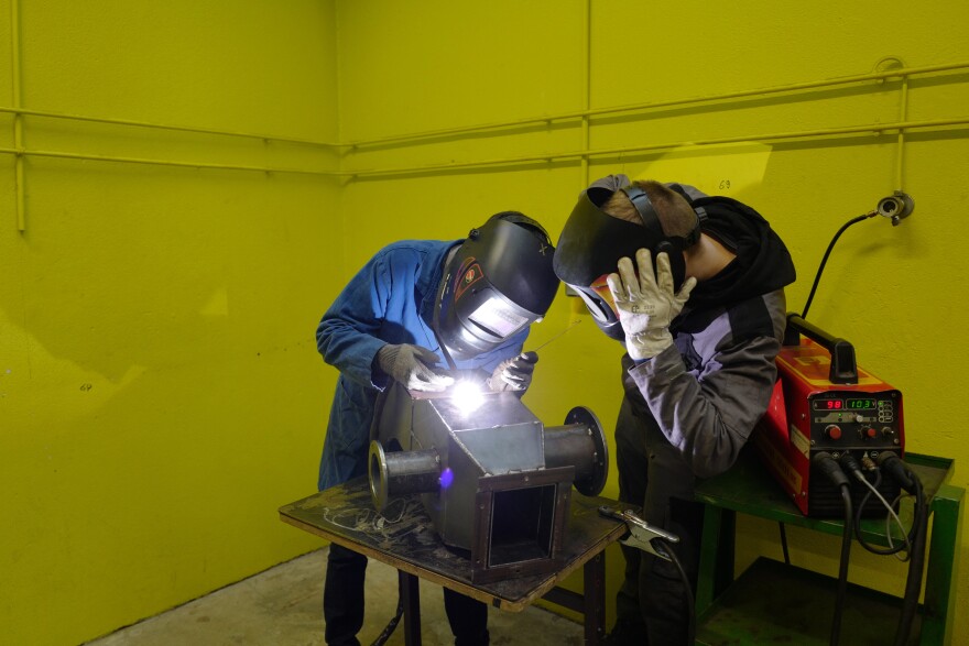 Two people wearing protective gear practice welding in an empty lime green room. 