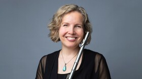 Flutist Lorna McGhee, holding her instrument and smiling at the viewer, wearing a black dress against a dark blue background
