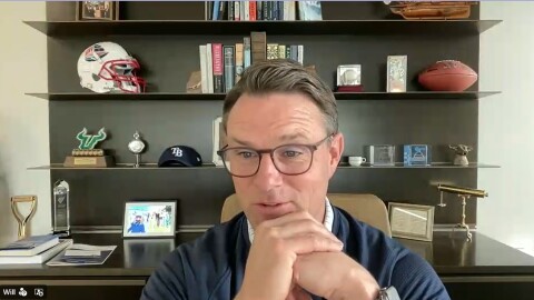 A man in glasses and navy sweater clasps hands, sitting in front of a bookcase with sports memorabilia and a dozen or so books