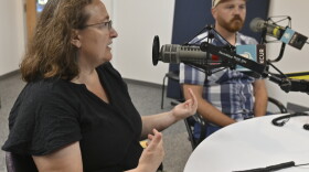 Two people sit in a radio studio talking at microphones. The person at left is gesturing while talking.