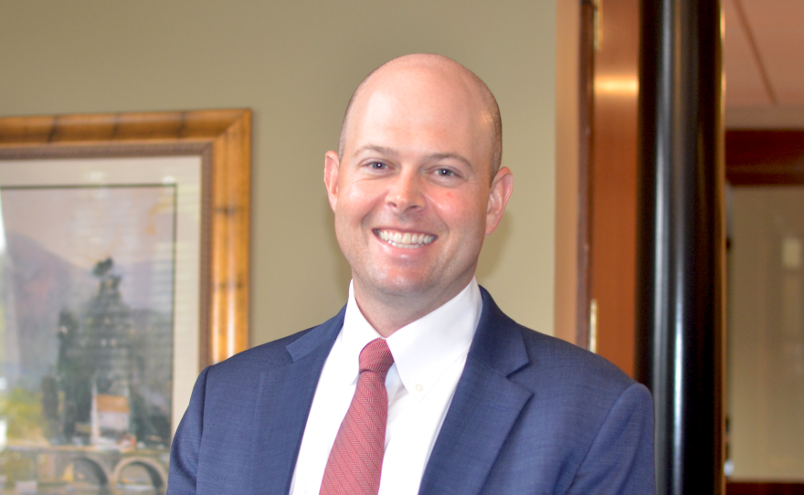 A smiling bald man wearing a white button-down shirt, red striped tie, and a blue suit coat