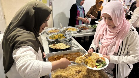 A woman is serving a dish of rice, chicken and lamb to another women who is holding a plate. There are trays of food between them. The women on the left is wearing a green headscarf and the women on the right is wearing a pink headscarf.