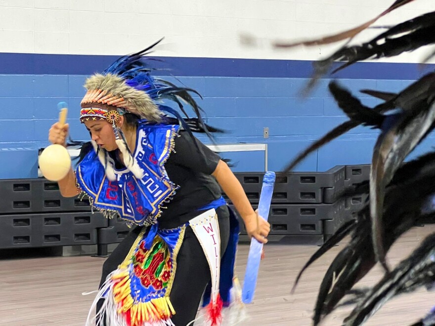 A dancer dressed in blue shakes a noise maker. She bends and moves to the beat of a drum.