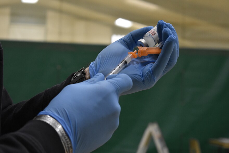 A close-up of latex-gloved hands pulling a syringe from a vial of Moderna COVID-19 vaccine.