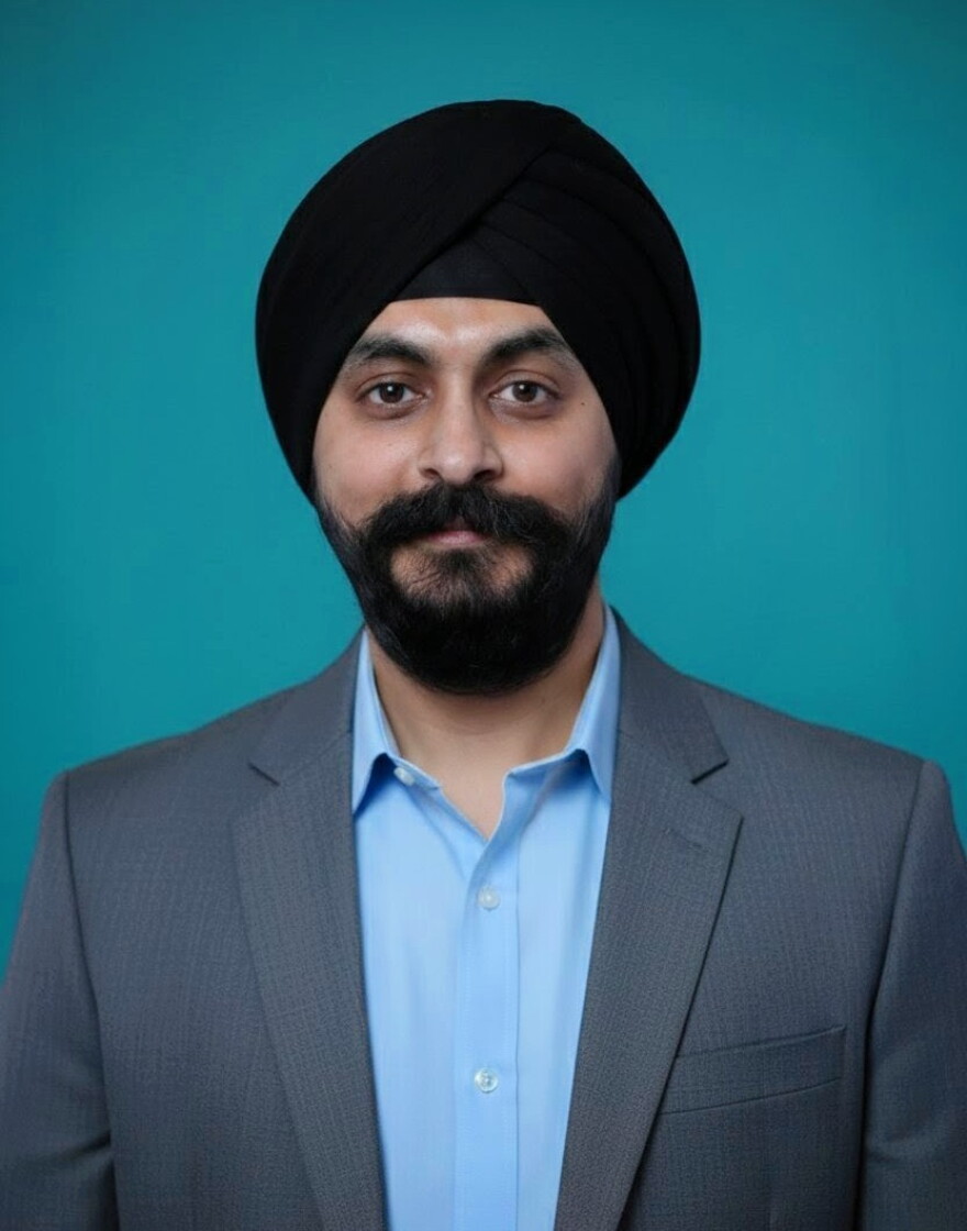 a portrait picture of a man in a blue shirt and grey blazer wearing a turban against a turquoise background